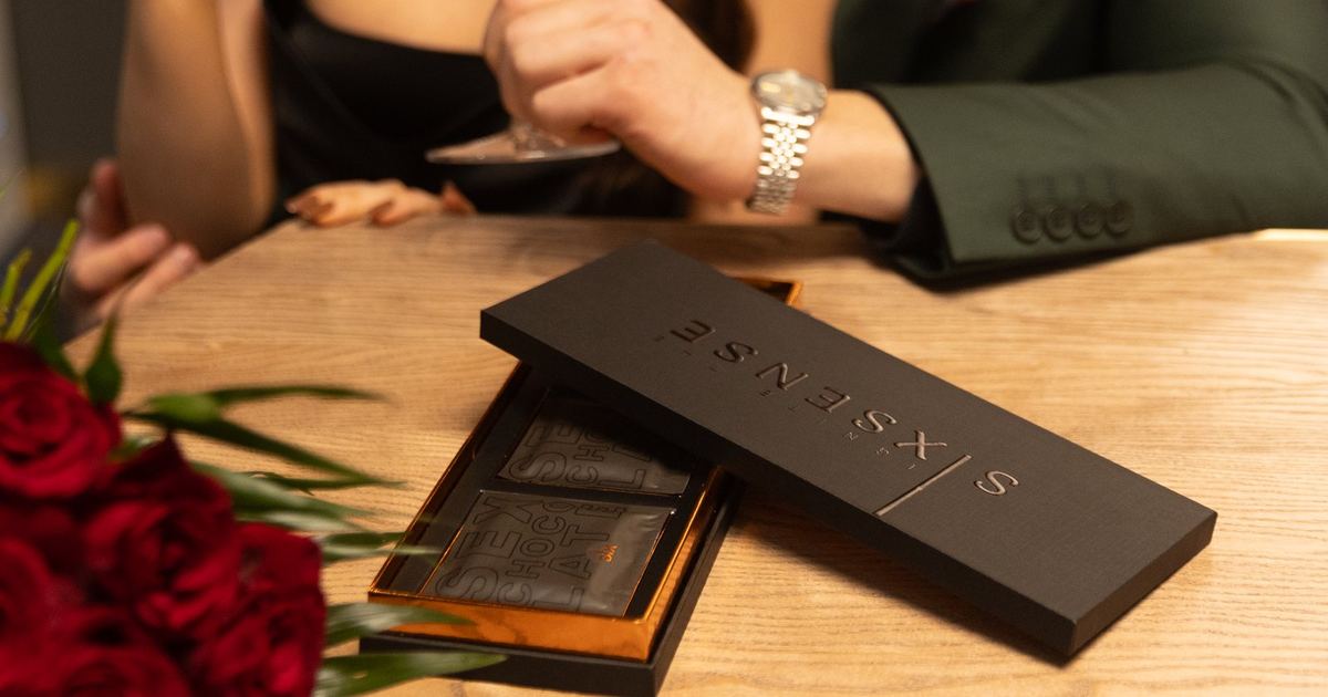 SIXSENSE couples chocolate box open on a table, showing individually wrapped dark chocolates, with red roses and a couple sharing wine in the background